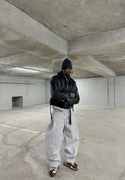 Hombre con gorro negro y chaqueta de cuero, con los brazos cruzados, se encuentra en un estacionamiento de concreto vacío, vistiendo pantalones deportivos grises y zapatos con estampado.