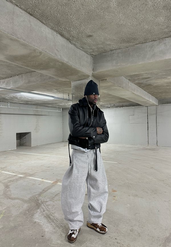 Man in een zwarte muts en leren jas staat met gekruiste armen in een lege betonnen parkeergarage, gekleed in grijze joggingbroek en patroon schoenen.