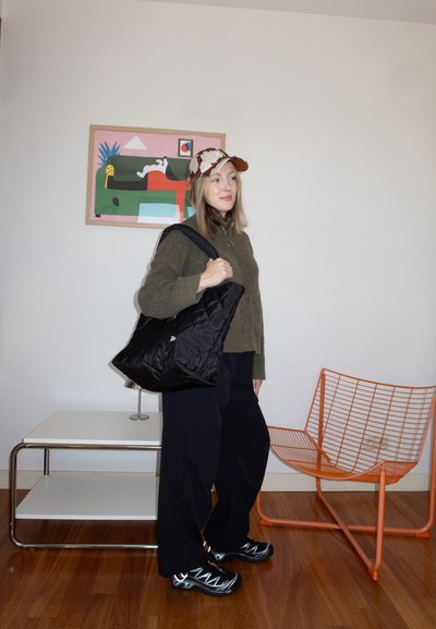Mujer con suéter verde, pantalones negros y gorra estampada, con un bolso negro grande, de pie junto a una silla de alambre naranja y arte abstracto en la pared.