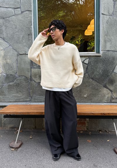 Suéter acanalado color blanco roto con hombros caídos, sobre una camisa blanca; pantalones negros de corte amplio y zapatos negros; entorno al aire libre.