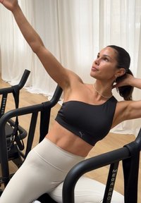 Černá sportovní podprsenka s jedním ramínkem v kombinaci s bílými legínami s vysok�ým pasem, na pozadí cvičebního vybavení a světlého pozadí.