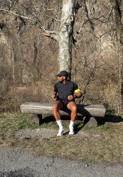 Corredor con ropa deportiva negra y gorra sentado en un banco de madera, mirando hacia un lado, con árboles sin hojas y terreno rocoso de fondo.