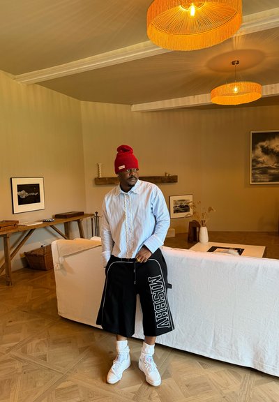 Hombre con gorro rojo, camisa azul claro, pantalones cortos negros con texto blanco, calcetines blancos y zapatos, de pie en una sala de estar beige con suelo de madera y lámparas naranjas.