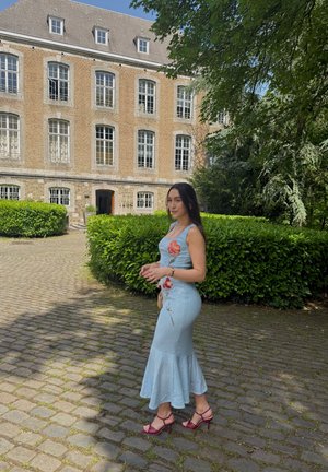 Mujer con un vestido azul claro con flores y tacones rojos caminando por un camino empedrado cerca de un edificio histórico de ladrillo con grandes ventanas y arbustos verdes.