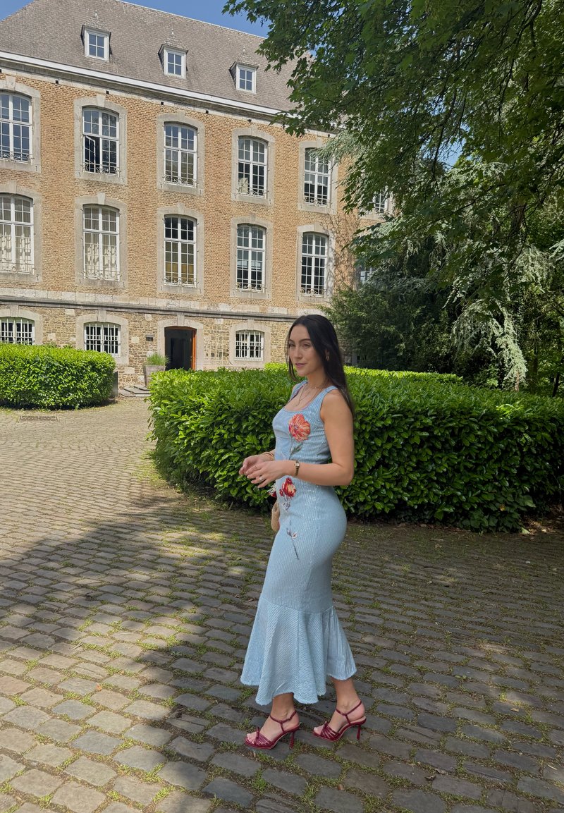 Femme en robe fleurie bleu clair et talons rouges marchant sur un chemin pavé près d'un bâtiment historique en briques avec de grandes fenêtres et des buissons verts.