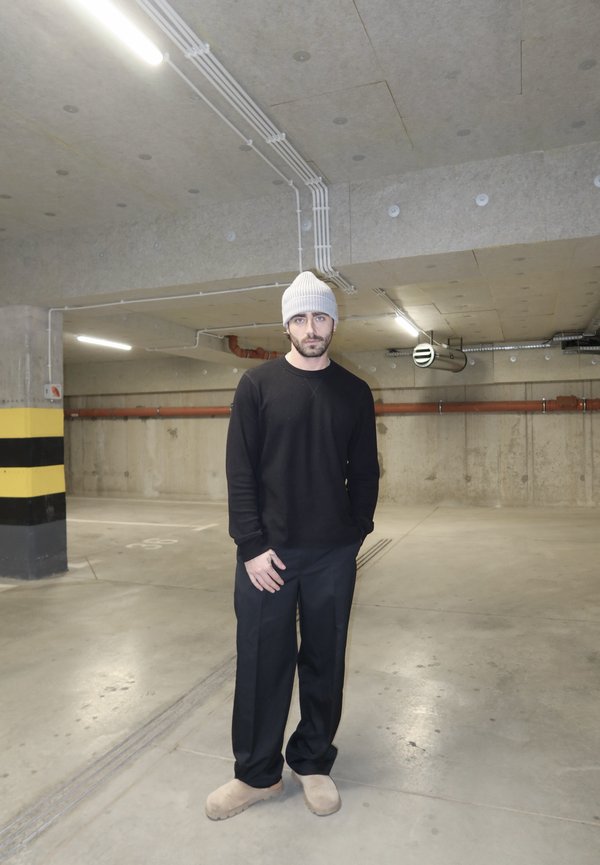 Mann trägt weiße Mütze, schwarzen Pullover, schwarze Hose und beige Schuhe, steht in einer leeren Beton-Tiefgarage.
