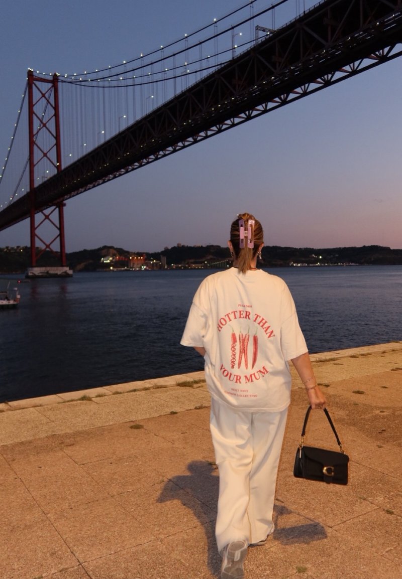 Λευκό oversized t-shirt με κόκκινο κείμενο και γραφικά; συνδυάζεται με χαλαρά λευκά παντελόνια. Τα αξεσουάρ περιλαμβάνουν μια μαύρη τσάντα και clips για τα μαλλιά.