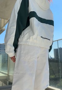 Giacca leggera bianca con accenti verdi, polsini elasticizzati e cappuccio regolabile, abbinata a pantaloncini di denim bianchi con tasche posteriori.
