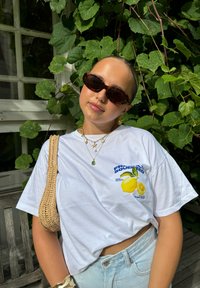 Witte cropped t-shirt met een citroenafbeelding; gecombineerd met lichte denim shorts, gelaagde gouden kettingen, tortoise zonnebril en een rieten tas.