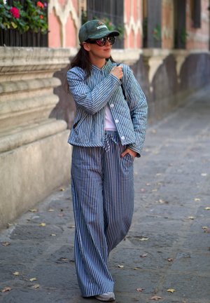 Chaqueta a rayas azul y pantalones de pierna ancha hechos de una tela ligera. Los accesorios incluyen una gorra verde y gafas de sol, con un ajuste casual.