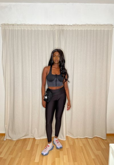 Top corto negro con cremallera frontal, leggings negros de cintura alta y zapatillas moradas claras con detalles rojos. Pequeño bolso cruzado negro.