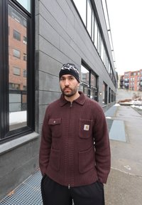 Homme avec barbe et bonnet noir portant un pull à fermeture éclair marron, debout à l'extérieur près d'un bâtiment gris avec de grandes fenêtres dans une zone urbaine.