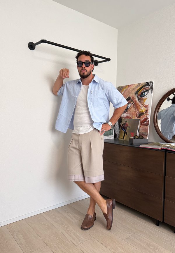 Mann mit Sonnenbrille, hellblauem Hemd, weissem T-Shirt, beigen Shorts und braunen Schuhen lehnt an der Wand neben einer Kommode mit Kunstwerken und rundem Spiegel.