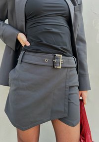 Γκρι μίνι φούστα  με διαγώνια στρίφωμα, θηλιές ζώνης και μαύρη τετράγωνη αγκράφα. Φορεμένο πάνω από ένα ταιριαστό τοπ, συνδυασμένο με μια κόκκινη τσάντα χειρός.