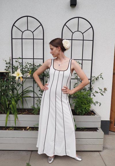 Vestido maxi blanco ajustado con Rayas verticales negras, escote cuadrado y tirantes finos. Zapatillas planas plateadas complementan el conjunto.