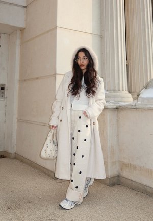 Mujer con abrigo largo blanco, pantalones de lunares y zapatillas de deporte, apoyada contra una pared de piedra beige con columnas, sosteniendo un bolso blanco con tachuelas.