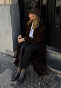 Femme en manteau marron et casquette assise sur une marche, tenant un sac à main marron, portant un pantalon à rayures fines et des chaussures marron sur un trottoir en ville.