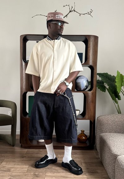 Hombre con camisa blanca de manga corta abotonada, pantalones cortos anchos oscuros con cadena, calcetines blancos, zapatos negros y gorra estampada, de pie en el interior junto a una estantería y plantas.