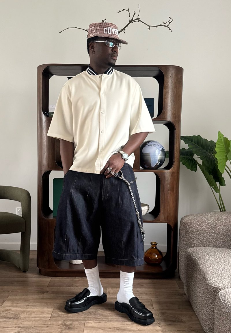 Homme portant une chemise blanche à manches courtes boutonnée, un short large foncé avec chaîne, des chaussettes blanches, des chaussures noires et une casquette à motifs, debout à l'intérieur près d'une étagère et de plantes.