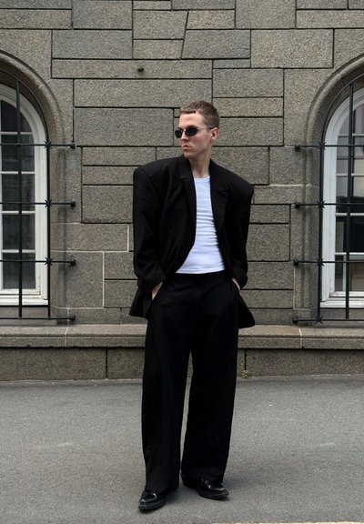 Blazer negro de gran tamaño con hombros exagerados sobre una camisa blanca y pantalones anchos negros. Unos zapatos negros elegantes y gafas de sol completan el look.