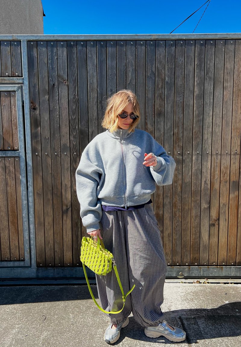 Person trägt übergroße graue Jacke mit Reissverschluss, karierte weite Hose, weiße Sneakers, Sonnenbrille, hält eine knallgrüne Handtasche, steht bei einem Holztor.