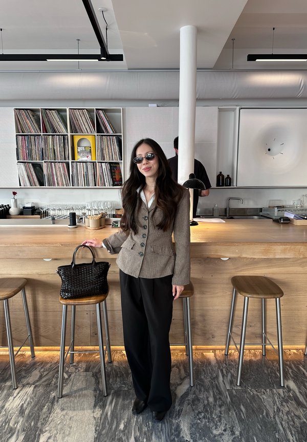 Una mujer con gafas de sol y una americana de cuadros está de pie junto a un bolso sobre un taburete, en la barra de una cafetería moderna llena de discos.