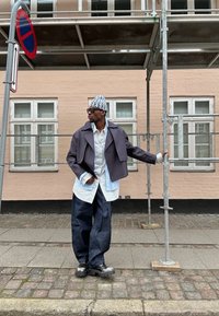 Henkilö, joka käyttää harmaata lyhyttä takkia, valkoista paitaa ja tummia farkkuja. Mustat kengät paksuilla pohjilla. Taustalla näkyy rakennus ja telineitä.