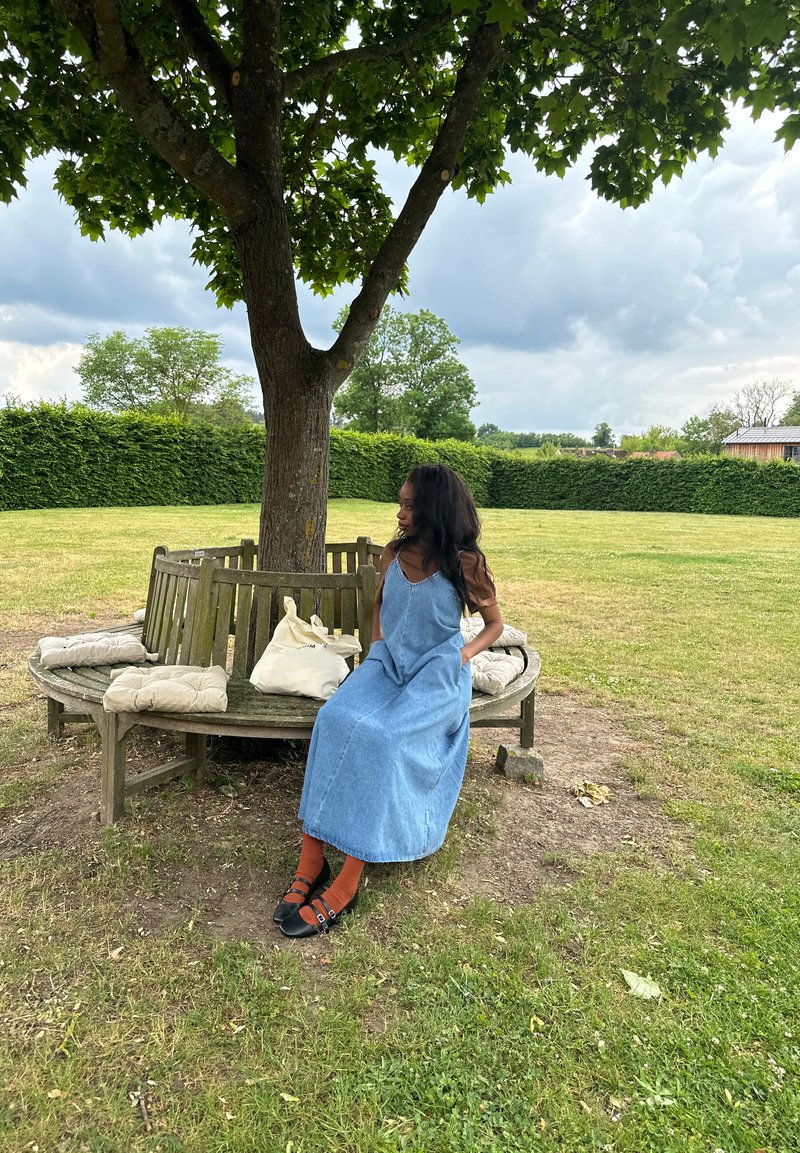 Une femme en robe bleue est assise sur un banc en bois rond autour d'un arbre dans un parc herbeux avec des coussins et un sac fourre-tout à proximité.