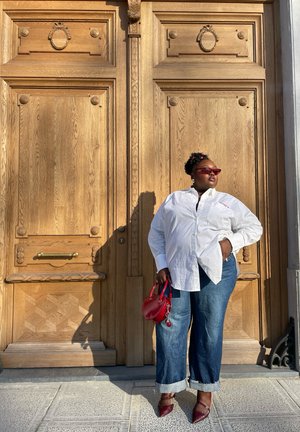 Camisa blanca de botones, jeans de mezclilla azules con los puños arremangados, bolso rojo y zapatos burdeos de punta, de pie frente a puertas de madera con detalles ornamentales.