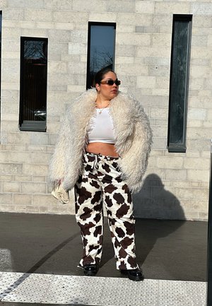 Abrigo blanco esponjoso y oversize, top corto blanco ajustado, pantalones de pata de elefante con estampado de vaca, y zapatos de plataforma negros; unas gafas de sol completan el look.