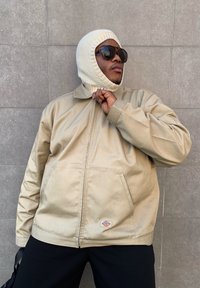 Veste zippée beige avec un col haut, dotée d'une capuche en tricot texturé et de lunettes de soleil sombres, devant un mur carrelé gris.