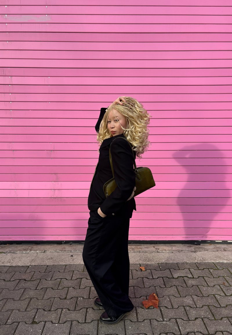 Zwart pak met wijde broek, draagt een groene tas. Lang golvend blond haar, roze achtergrond, leunt tegen een muur, schaduw valt op het trottoir.