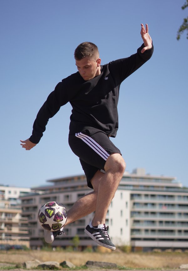 Joven con ropa deportiva negra saltando en el aire mientras controla un balón de fútbol, con edificios urbanos al fondo bajo cielo despejado.