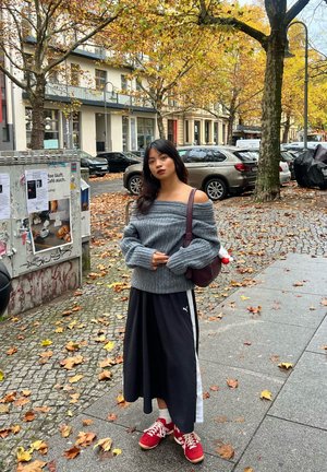 Suéter gris de gran tamaño con diseño off-shoulder, falda negra con raya blanca, zapatillas rojas y un bolso marrón en una calle de adoquines entre hojas de otoño.