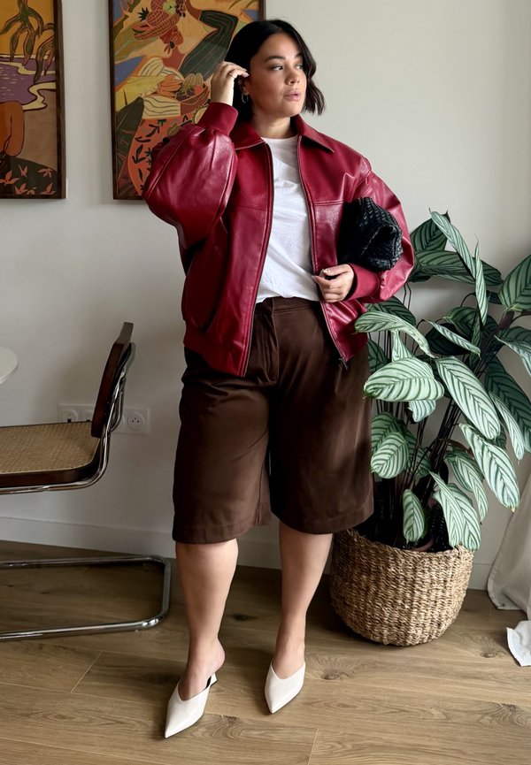 Une femme ajuste ses cheveux tout en posant à côté d’une plante en pot, vêtue d’une veste en cuir rouge, d’un haut blanc, d’un short brun et de chaussures à talons blanches.
