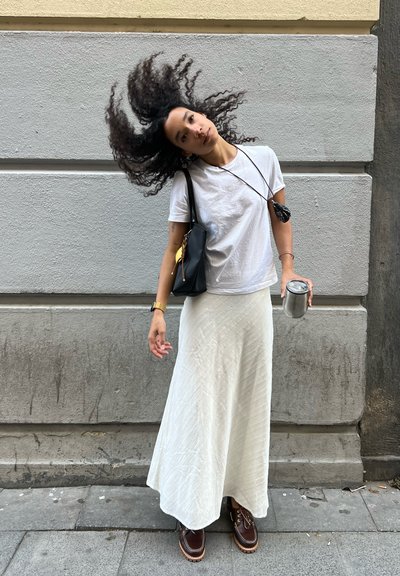 Mujer con cabello rizado moviendo la cabeza, vestida con camiseta blanca y falda larga, sosteniendo una taza de metal y un bolso negro, de pie contra una pared gris.