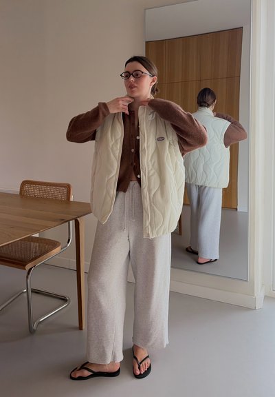 Mujer ajustándose un chaleco acolchado crema sobre un suéter marrón, con pantalones anchos grises y sandalias negras, de pie cerca de una mesa de madera y un espejo.