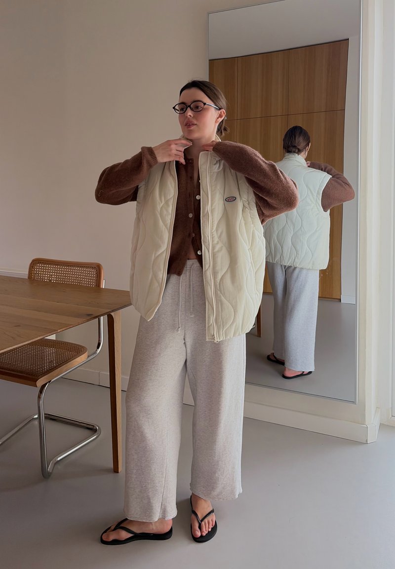 Femme ajustant un gilet matelassé crème par-dessus un pull marron, portant un pantalon large gris et des tongs noires, debout près d'une table en bois et d'un miroir.