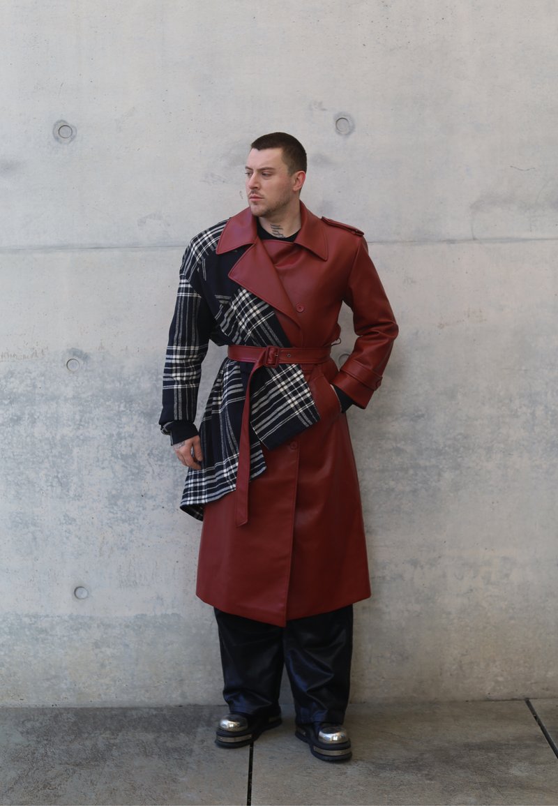 Trench-coat en similicuir marron avec une manche en tissu écossais, taille ceinturée et pantalon large noir ; des chaussures métalliques brillantes complètent le look.