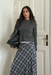 Femme debout à l'intérieur près d'une porte blanche, portant un cardigan gris, une jupe à carreaux, tenant un grand sac en cuir noir sous le bras gauche.