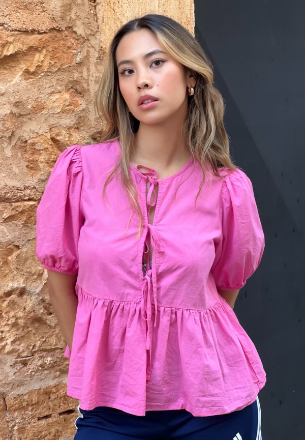 Pink, puff-sleeve blouse with a drawstring tie at the neckline. Features a flared hem and lightweight fabric, displayed against a stone wall.