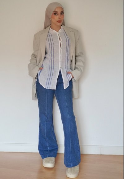 Blazer gris claro sobre una camisa blanca de botones con rayas azules, combinado con jeans acampanados azules y zapatos de plataforma beige. Fondo liso.