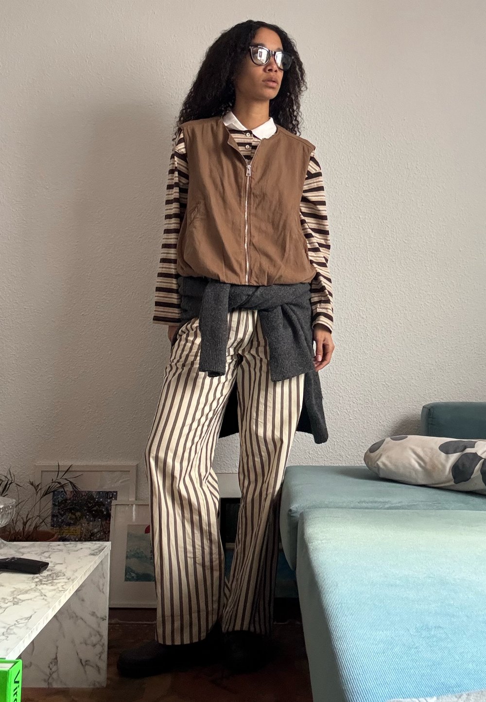 Femme aux cheveux bouclés portant des lunettes de soleil, un gilet marron sans manches, une chemise et un pantalon rayés, avec un pull gris noué autour de la taille à l'intérieur.