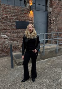 Jaqueta de couro preta sobre um top preto canelado, calças pretas largas e botas pretas de bico fino. Cabelo loiro longo estilizado em caracóis, apoiada contra uma parede de tijolos.