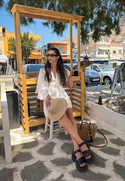 Vestido blanco texturizado con mangas acampanadas, combinado con sandalias de plataforma negras. Adornado con un sombrero de paja y un bolso de mano trenzado en marrón. Entorno al aire libre.