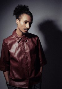 Chemise en cuir bordeaux avec un col pointu, des manches courtes et un devant à boutons. Texture lisse avec un léger éclat. Fond ombragé.