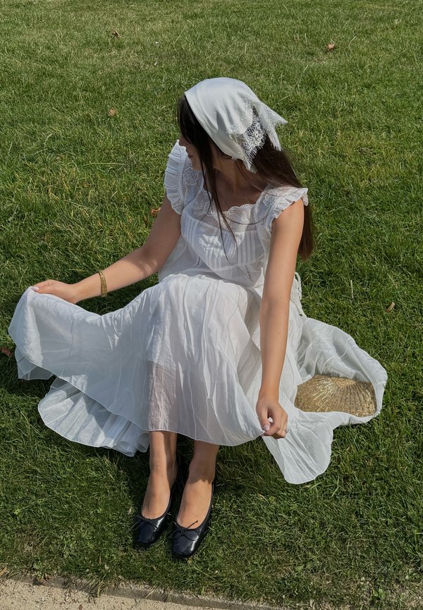 Femme en robe blanche et foulard sur la tête assise sur l'herbe, tenant sa jupe, avec un chapeau de paille à côté d'elle et portant des ballerines noires.