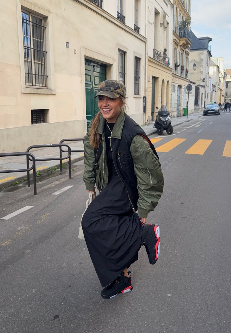 Femme portant une veste verte et noire, une robe noire, une casquette camouflage et des baskets noires et rouges, levant un pied dans une rue de la ville avec des bâtiments et une trottinette en arrière-plan.
