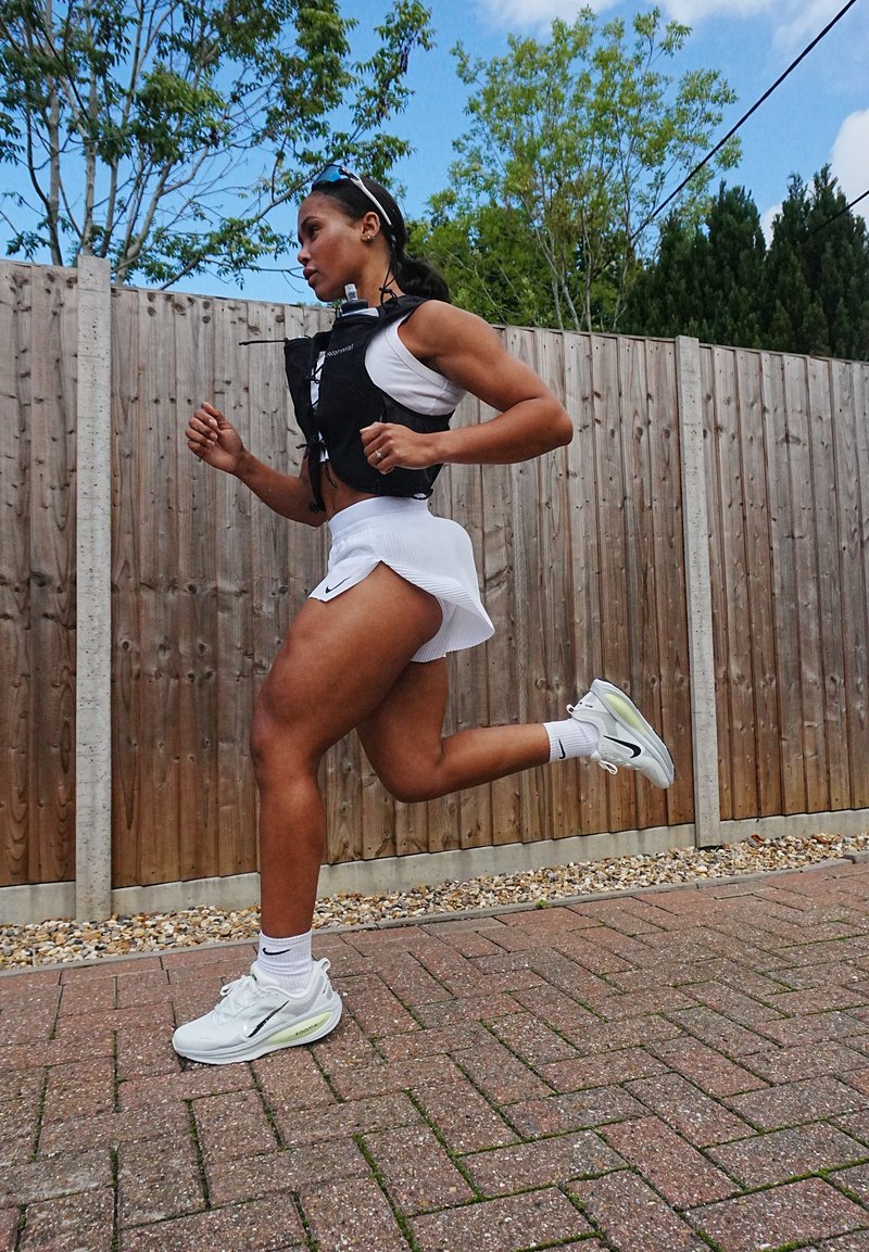 O alergător îmbrăcat cu un vestă neagră peste un top alb și o fustă, purtând adidași albi și gri, cu un fundal de asfalt texturat și gard din lemn.