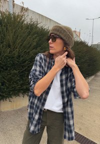 Cămașă în carouri bleumarin și alb, tricou alb dedesubt, pantaloni verde măsliniu, căciulă bej olive și ochelari de soare închiși la culoare; fundal în tonuri pământii.